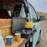 Van with a portable stove, yellow kettle, and floral mugs on a wooden shelf.
